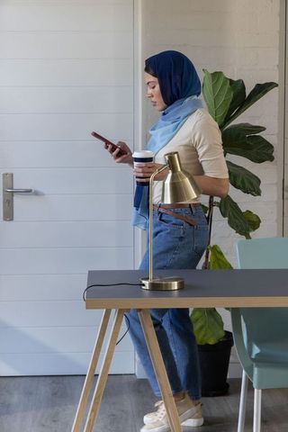 Young woman wearing navy hijab using smartphone and coffee at minimalist home workspace