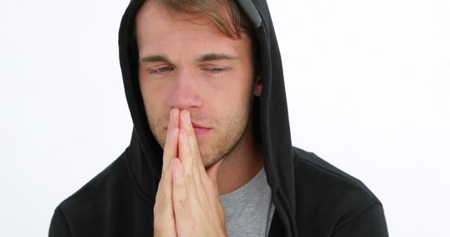 Young man wearing black hoodie and grey shirt praying with serious expression. Perfect for concepts of prayer, devotion, focus, introspection, or concern. Can be used in religious articles, motivational posters, or mindfulness promotion material.