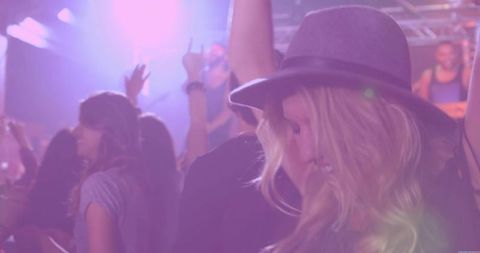 Dancing blonde woman raising arm under purple neon lights wearing fedora at nightclub