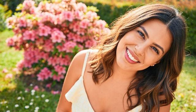 Sunlit smiling woman in yellow sundress leaning toward camera in blooming garden