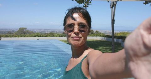 Mature woman taking selfie at pool edge wearing green swimsuit and sunglasses, backyard view