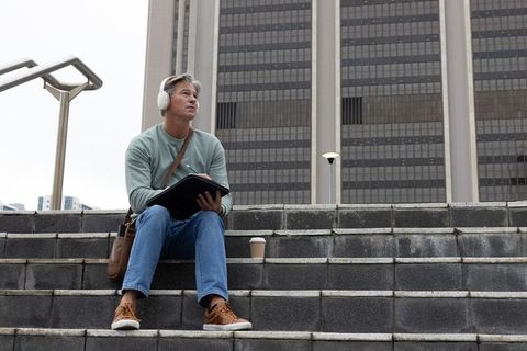 Middle-aged man sitting on urban steps taking notes with headphones and coffee cup