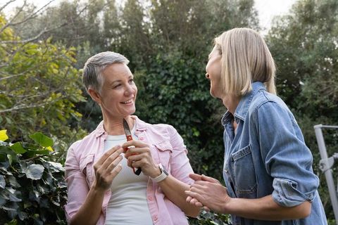 Mature woman pruning hedge with friend laughing in sunny backyard garden casual teamwork