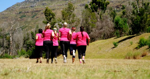 Group of women wearing pink shirts running in an outdoor natural setting. Green trees and hills create a scenic background. Ideal for use in fitness campaigns, health and wellness promotions, or running event advertisements.