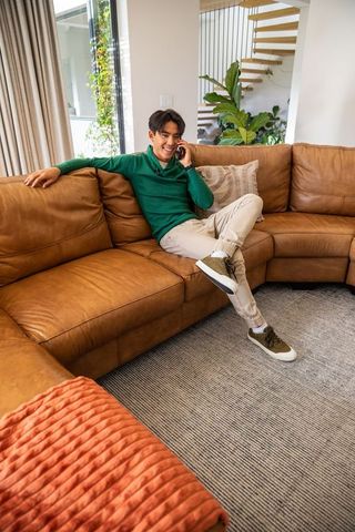 Young asian man relaxing on tan leather sectional talking on smartphone in living room