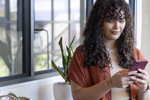 Young woman using smartphone by window in bright office with plants and casual style