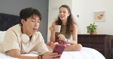 Young couple relaxing on bed using smartphone and drinking coffee in minimalist bedroom