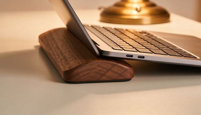 Curved walnut wrist rest supporting slim laptop with visible usb-c ports, warm desk light