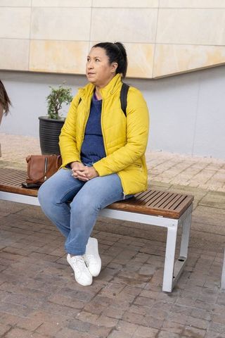 Mature woman in yellow jacket sitting on bench, glancing at friend in urban courtyard