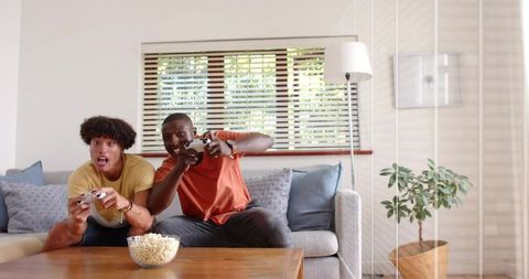 Two friends playing competitive video game on couch, laughing with gamepads and popcorn