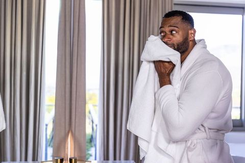 Mature african american man in bathrobe drying face with towel by sunlit window and balcony