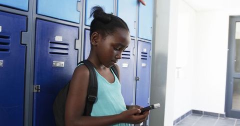African female teen checking smartphone at blue lockers wearing teal dress and backpack