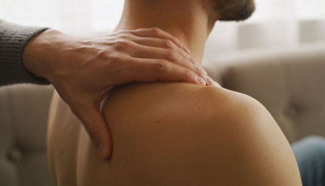 Hand massaging shirtless male shoulder and upper back on tufted sofa in warm daylight