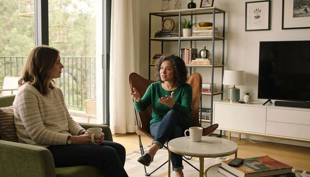 Two women having heartfelt conversation in cozy living room with coffee and balcony view