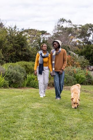 African american couple holding hands walking through autumn garden with tan dog