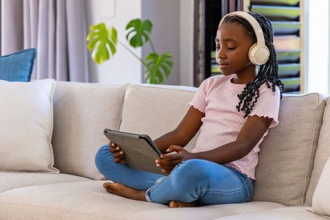 Girl wearing headphones using tablet on living room sofa for home learning and focus