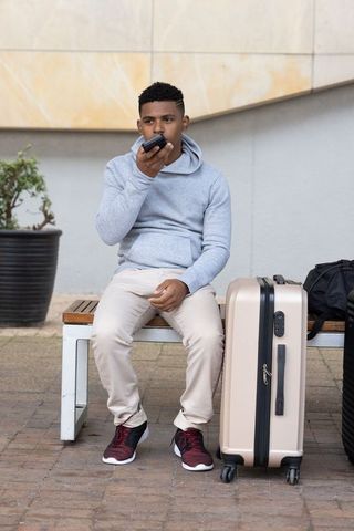 African-american traveler waiting on bench holding phone with suitcase and backpack
