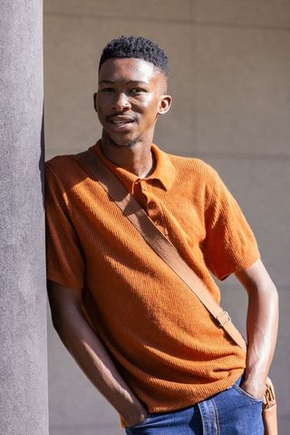 African man leaning on pillar wearing orange polo and crossbody bag, sunlit urban portrait