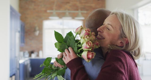 Senior couple sharing a happy moment in a cozy kitchen, holding a bouquet of roses. Suitable for themes of romance among mature adults, joyful relationships, and domestic happiness. Excellent for use in advertisements focused on love, elder care, and lifestyle products targeting mature audiences.