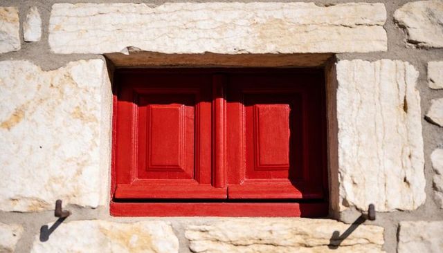 Sunlit red wooden shutters in limestone facade with rustic iron hooks and texture