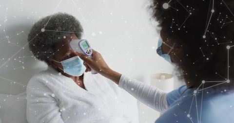 Home caregiver taking senior's forehead temperature with infrared thermometer, wearing masks