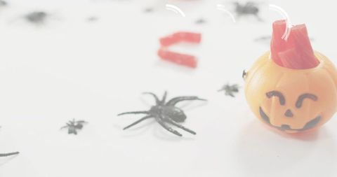 Smiling jack-o'-lantern sitting among plastic spiders and red candy on white surface
