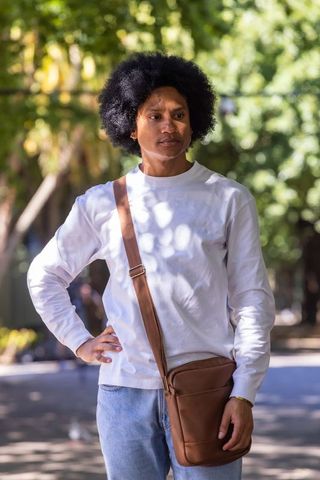 Standing man wearing white long sleeve and brown crossbody bag on sunlit tree-lined path