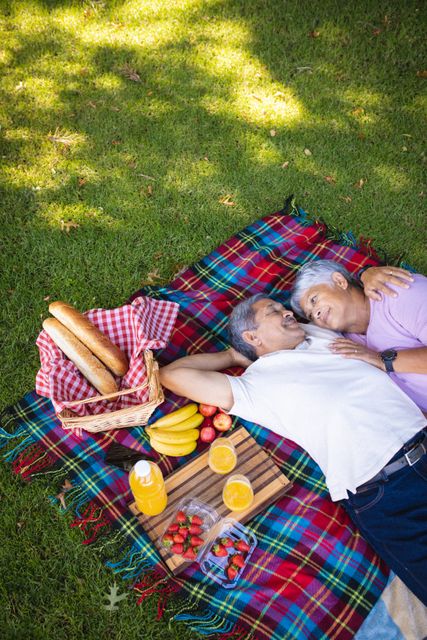 High angle view of biracial senior couple looking at each other while lying on blanket in park. romance, food, drink, picnic, nature, unaltered, love, togetherness, lifestyle and retirement concept.