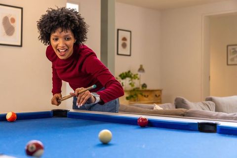 African american woman playing pool at home on blue-felt table in cozy living room