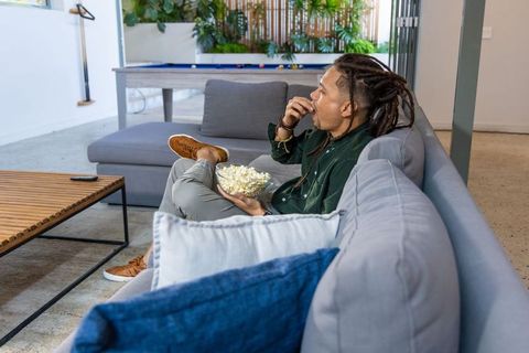 Young african american man relaxing on modern sectional, snacking on popcorn in stylish lounge