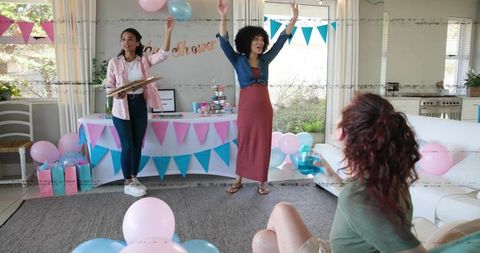 Joyful expectant mother raising arms at pastel baby shower with friends and festive decor