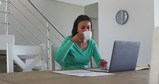 Woman in casual dress working remotely on laptop in bright, modern home while drinking coffee. Papers on table hinting at business or academic tasks. Can be used for promoting remote work, home office setups, casual productivity, and real estate interiors.