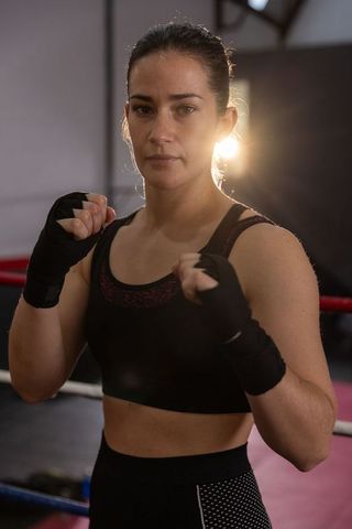 Female boxer standing in ring holding wrapped fists backlit by warm gym light, focused