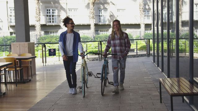 Two male friends walking side by side with bicycles in an urban environment. One wears a plaid shirt, while the other sports a jacket and sunglasses. Palm trees and large building in the background create a city park feel. Perfect for themes of green urban living, friendship, outdoor activities, and sustainable transportation.