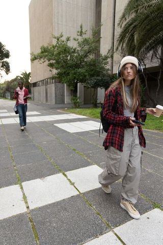 Young student walking university campus with headphones coffee smartphone backpack urban walkway