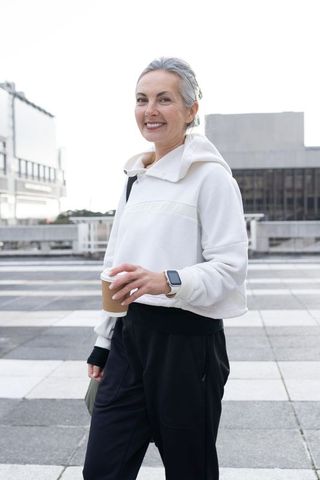 Active senior woman walking rooftop plaza holding coffee wearing hoodie and smartwatch