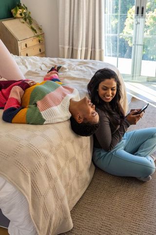 Sunlit bedroom friends sharing smartphone while lounging in colorful striped knit sweater