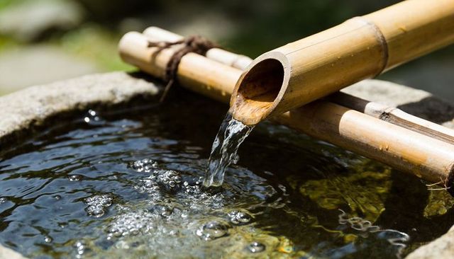 Bamboo spout flowing into stone basin creating ripples and sunlit reflections in garden