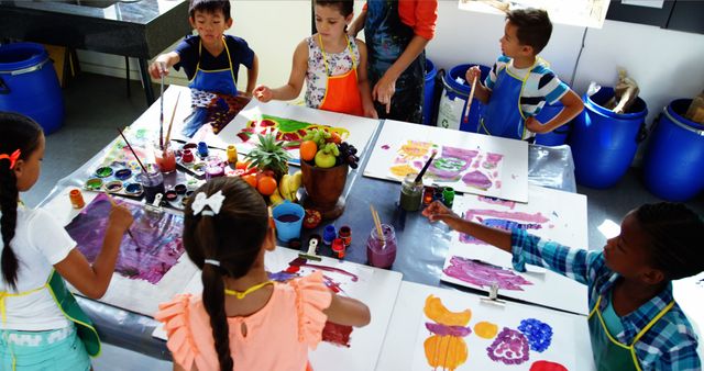 A group of diverse children is painting colorful artwork at a table in an art class. They use various colors and techniques, expressing their creativity. Ideal for themes related to childhood education, arts and crafts, diversity in schools, and collaborative learning activities.