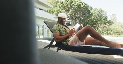 Mature man relaxing poolside on sun lounger reading book while wearing straw hat