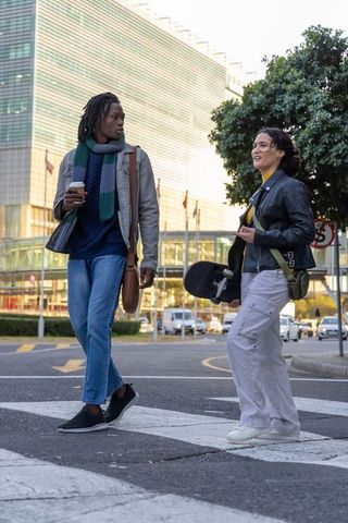 Young friends walking across urban crosswalk carrying skateboard coffee and satchel