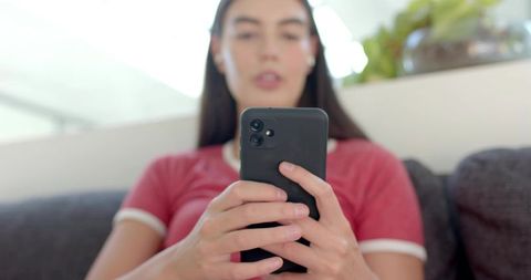 Young woman scrolling smartphone on sofa wearing earbuds and red ringer t-shirt