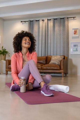 Smiling african american woman relaxing on yoga mat at home holding shaker bottle after workout