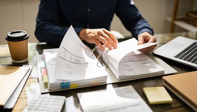 Business professional sorting invoices in binder on sunlit desk with laptop and coffee