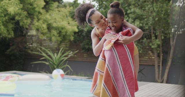 Depicts a mother wrapping her daughter in a towel after swimming by the poolside. It captures family bonding time during summer with an outdoor setting. This image can be used to illustrate concepts related to family, parenthood, summer fun, and outdoor activities. Brands promoting family products, summer vacations, or outdoor apparel may find this relevant.