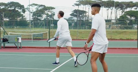 Male tennis teammates practicing doubles on outdoor hard court wearing white uniforms