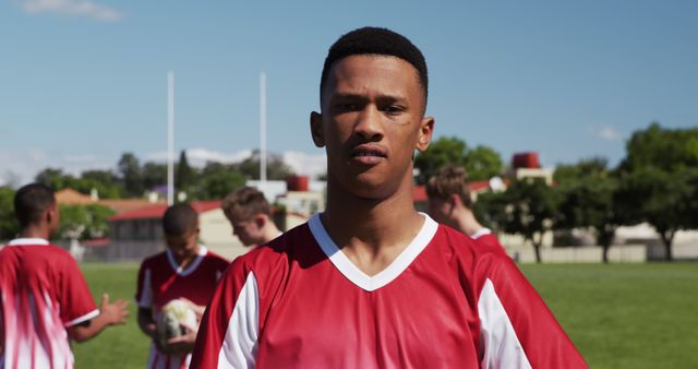 Teenage boy wearing a red jersey standing confidently on a soccer field with teammates and a ball in the background. Perfect for sports magazines, team sports promotions, active lifestyle campaigns, youth soccer programs, and exercise initiatives. Conveys teamwork, sportsmanship, and the energy of outdoor activities.