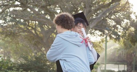 Diverse male friends hugging, celebrating graduation with cap and diploma outdoors