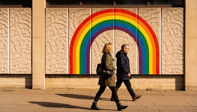 Walking past rainbow mural on textured concrete facade, urban pedestrians in coats