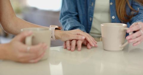 Two women holding hands over coffee mugs on glossy table conveying warmth and support